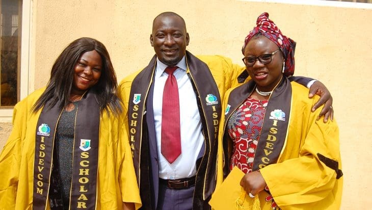 Three students from Nasarawa State University, dressed in bright yellows gowns, stand together smiling at the camera